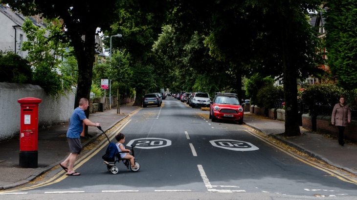 A parent pushing a pram across a residential 20mph street.

Rhiant yn gwthio pram ar draws stryd preswyl 20mya.