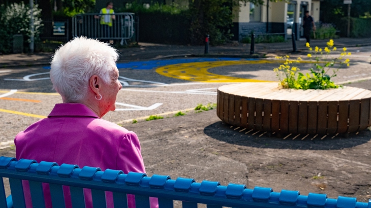 Elderly woman sitting on a bench surrounded by artworks and planters in Leith.