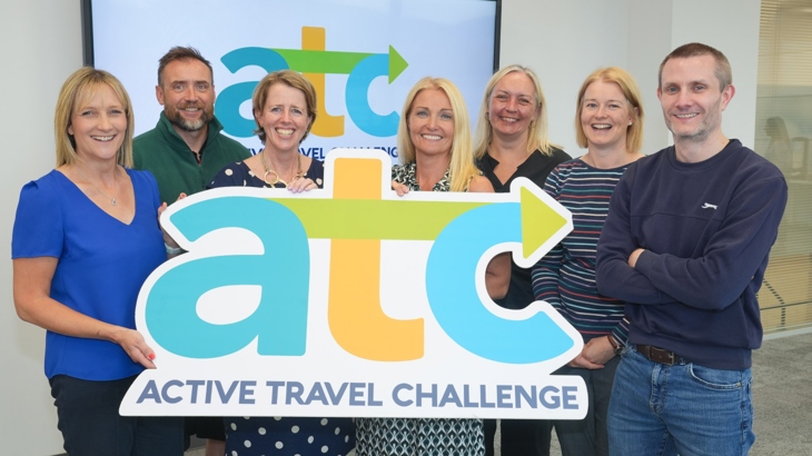 A group of smiling men and women stand indoors looking at the camera while holding a sign reading a-t-c.