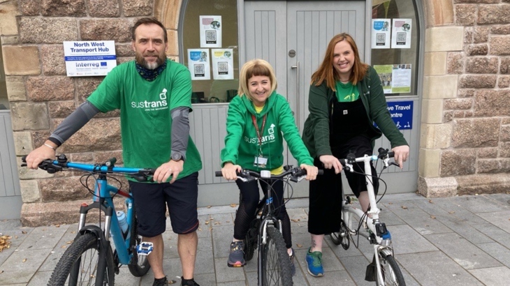 A man and two women stand with bikes outside an active travel centre wearing green Walk Wheel Cycle Trust branded clothing.