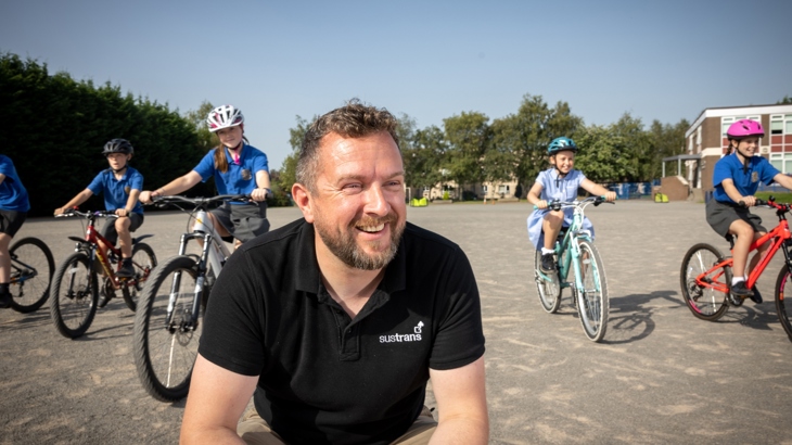 A man wearing a black Walk Wheel Cycle Trust top smiles at something off camera with children on bikes behind him in a school playground.