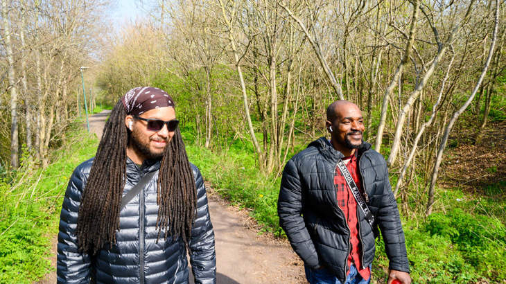 Two men chatting together as they walk along a traffic-free National Cycle Network route in Beckton, London surrounded by green grass and trees.