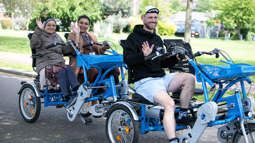A group of people smiling and waving as they cycle by on adapted cycles through a park.
