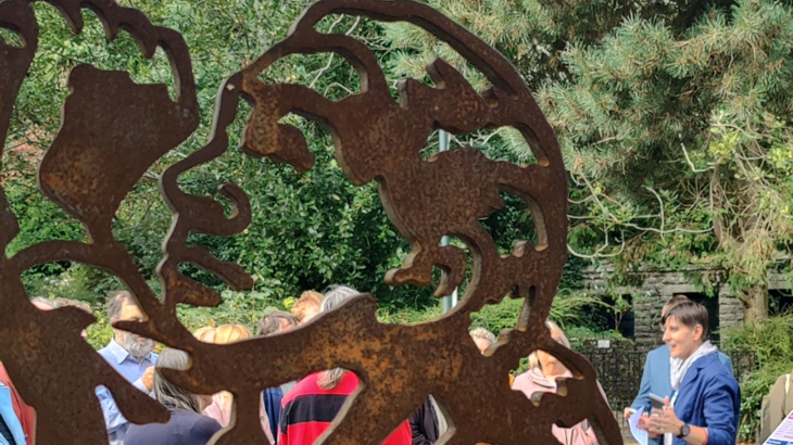 close up of steel portrait of Dr Goodall looking up to the trees with binoculars. People gather behind it talking. Trees line the background.
