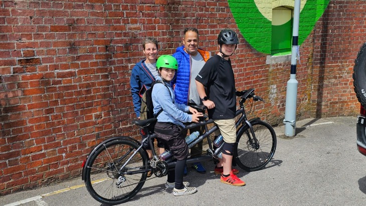 Paul Luttrell stands with his wife and two youngest sons as they sit on a tandem bike together in the sunshine.