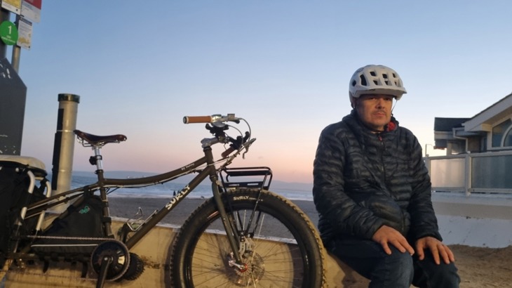Paul Luttrell sits on a wall next to his bike on a coastal path with a dusky sky above.