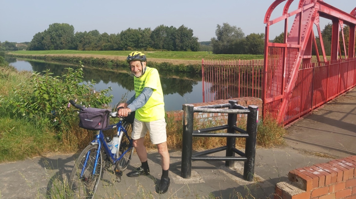 An older man with a high vis top on stood in the sunshine with his bicycle on a cycle route in the North of England