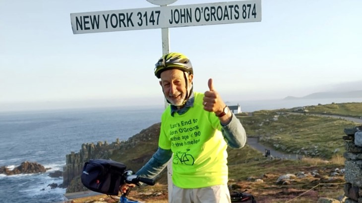 An older man wearing a bike helmet and a high vis top giving a thumbs up and smiling at a signpost at Land's End in Cornwall