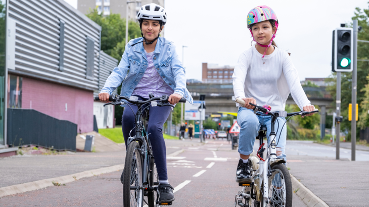 Two teenage girls on bikes wearing helmets cycling down a dedicated cycle lane in Glasgow.