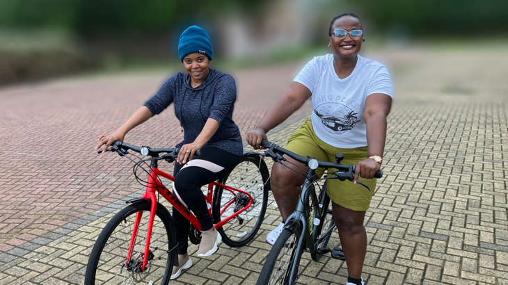 Two women sat stationary on their bikes, smiling. One is wearing a blue jumper and beanie hat, the other is wearing a white t-shirt and green shorts