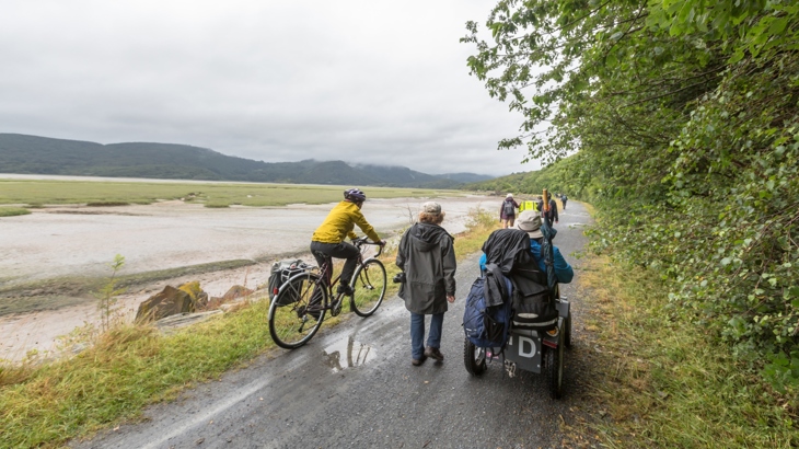 A cyclist, walker, and person using a mobility scooter travelling a scenic stretch of the National Cycle Network in Wales.