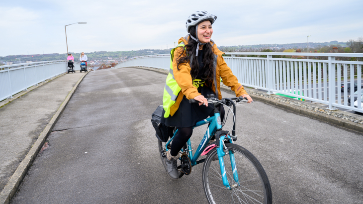 A young woman wearing a yellow raincoat and a helmet riding a bike on a flyover in an urban setting in England