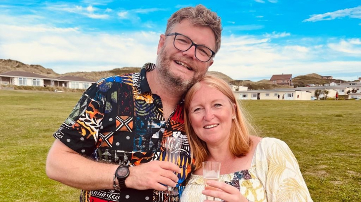 A man and a woman stood close together smiling on a grassy field holding a glass of alcohol each with blue skies in the background