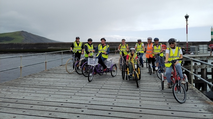 A group of adults on adaptive cycles in a rural setting, beside a body of water and with a hill in the background.