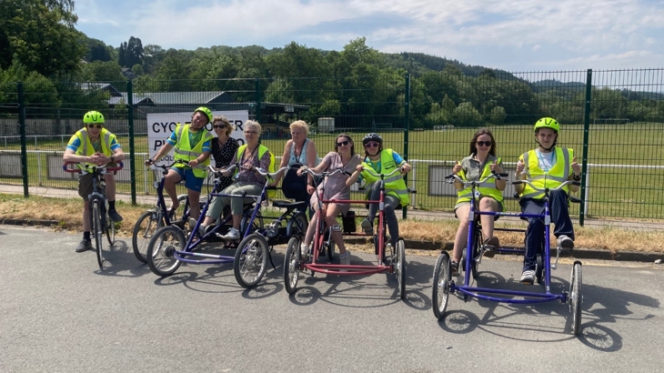 A group of adults posing on adaptive tandems and bicycles in front of a park area.