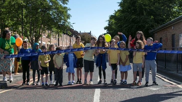 a group of children stand on a road in front of a ribbon, while one cuts it
