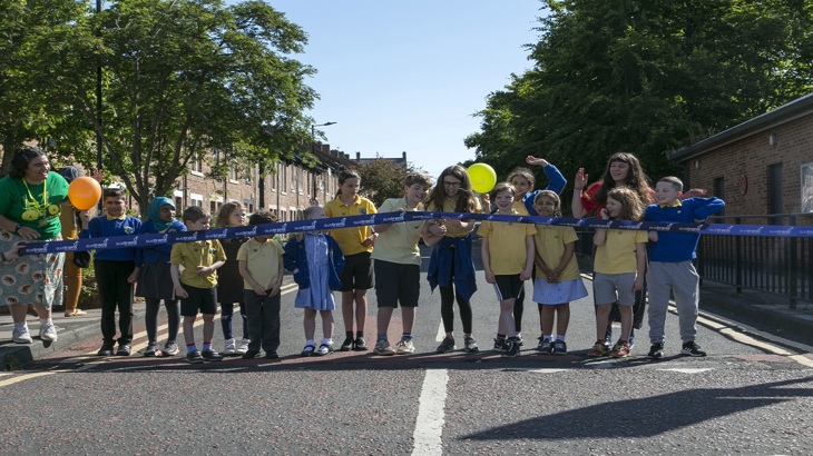 children stand on road behind a large ribbon. One is about to cut.