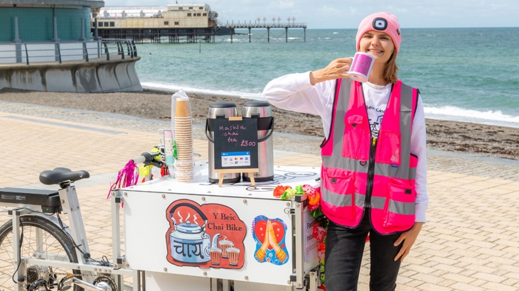 Sally standing on Aberystwyth promenade beside her XYZ e-cargo cycle holding up a cup of Masala chai.