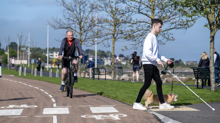 A young man with a visual aid crosses a cycle lane, with a cyclist approaching.