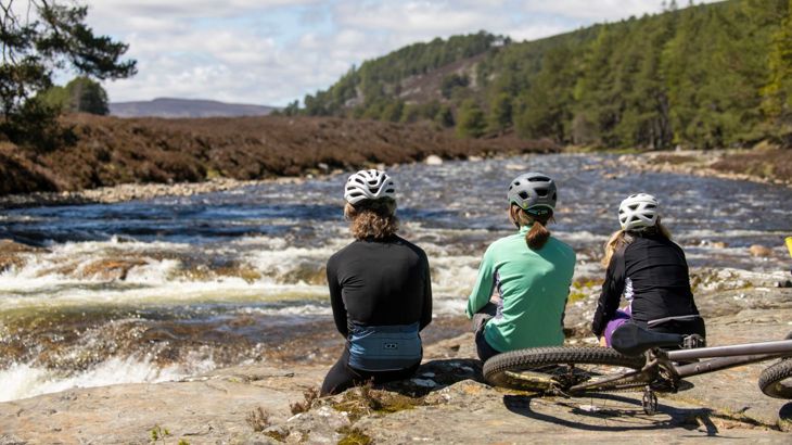 Three women sit by the water in the Cairngorms National Park, on the Lochs and Glens Way, with their bikes behind them