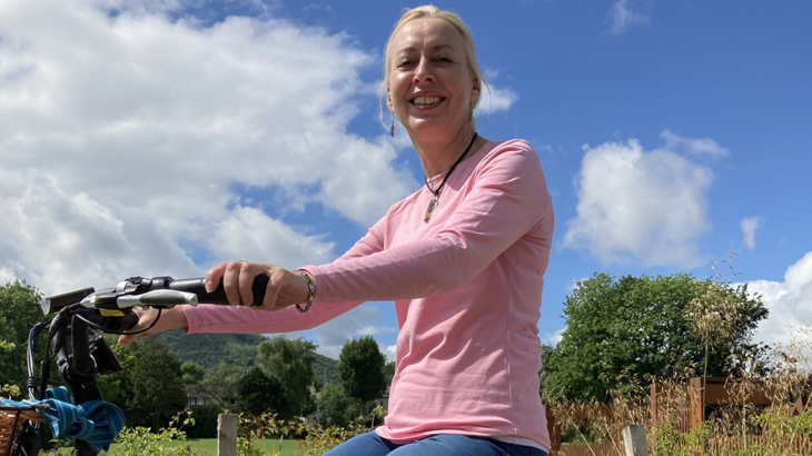 A woman wearing a pink top smiling in the sunshine while sat on her trike