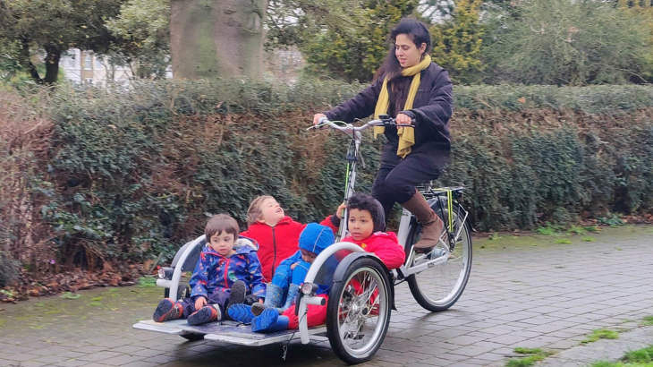 A woman rides an adapted cycle with a trailer on the front, in which several young children sit.