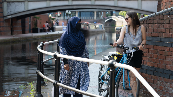 Two young women stood on a bridge smiling and chatting one is pushing a bike and the other is walking and wearing a blue hijab