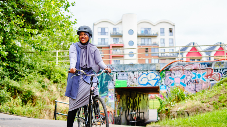 A woman smiling wearing a hijab on a bike near a graffitied underpass in a city on a bright day