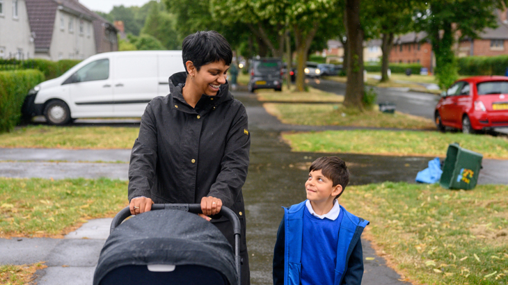 A parent pushing a pram in a residential area on a rainy day looking down at her child who is wearing a blue uniform and smiling looking up at her