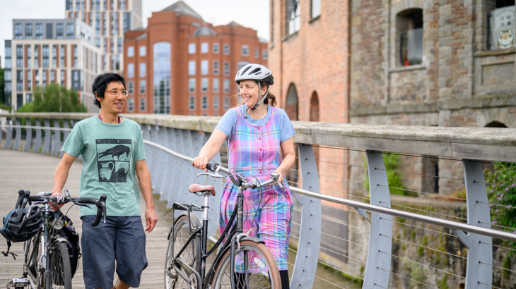 Two people walking along a bridge in a city both pushing their cycles along while chatting and smiling at each other on a cloudy day