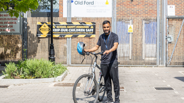 A male school teacher stood smiling outside school gates holding the handle bars of a bike with a blue helmet in one hand wearing sports clothing