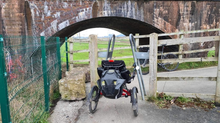 A recumbent trike is positioned in front of an A-frame barrier on a cycle path, demonstrating how it's unable to pass through.