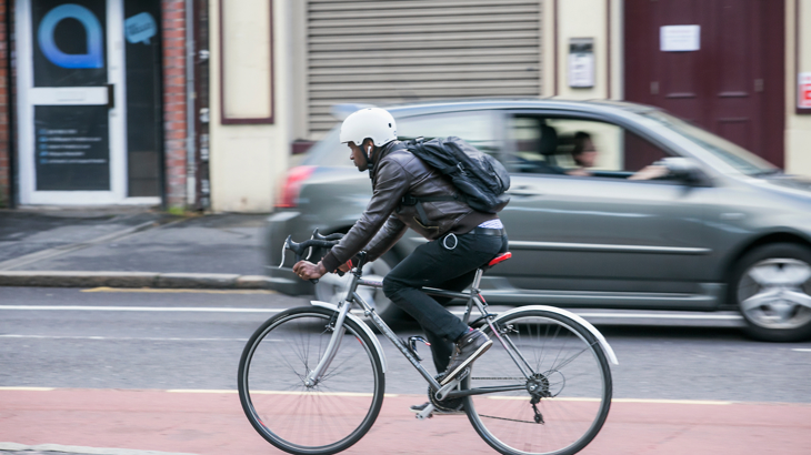 A man riding a bicycle down the street with a car passing nearby.