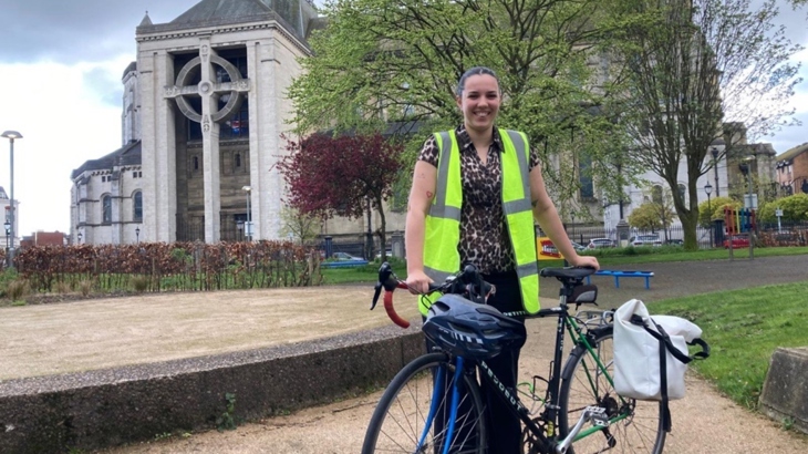 A woman wearing a hi-vis vest stands with a bike in a square in front of a cathedral.