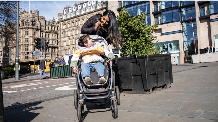 A mother is shown walking with her child in a pram in a busy city centre