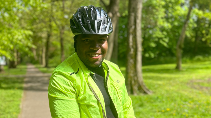 A man stood wearing high vis and a helmet smiling with grass and trees in the background on a sunny Spring day