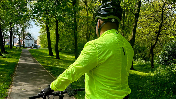 A man stood on his cycle wearing a high vis top and a black helmet facing away from the camera, looking down a traffic-free path lined with trees