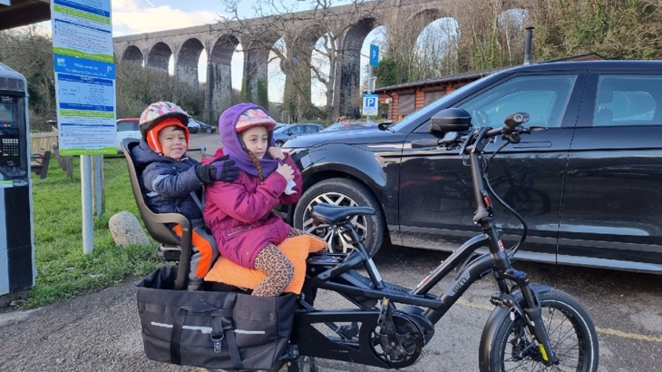 Two young children sat in child seats on the back of a black e-bike, both wearing helmets, which is parked in Porthkerry Park.