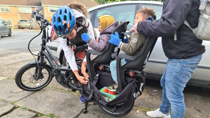 A mother is securing her two young children into their child seats on the back of a black e-bike, parked in a residential street.