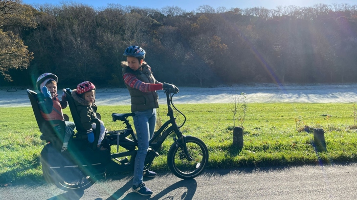 A mother and two young children are riding on a black e-bike in a park. All of them are wearing helmets. There's a mist on the grass in the background