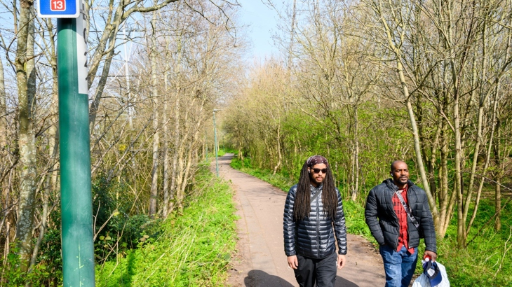 Two men walk along a tree-lined path in the sun, with a National Cycle Network sign in the foreground.