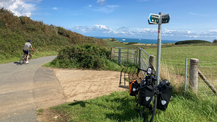 A cycle with panniers on either ride of it resting against a signpost on the side of a country lane on a sunny day with views of fields and the sea