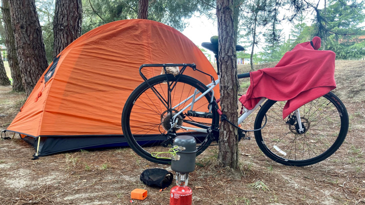 A white cycle propped up again a pine tree in a woodland setting next to an orange tent and a gas stove on the ground