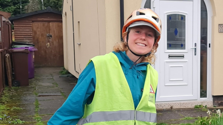 A woman with blonde curly hair stood smiling outside of a detached home wearing a high vis vest, a blue ran jacket and a helmet and a cap
