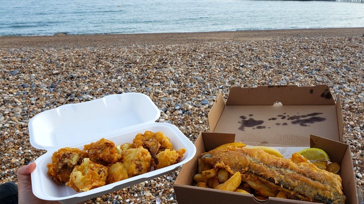 A box of fish and chips held up outside on Brighton beach on a cloudy day with the sea in the background