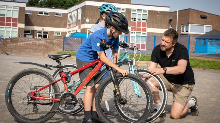 A man kneels beside two children on bikes to check their tyres in a school playground.