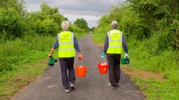 Two Walk Wheel Cycle Trust volunteers wearing high visibility jackets walking along a path holding red buckets with wildflower seedings in.