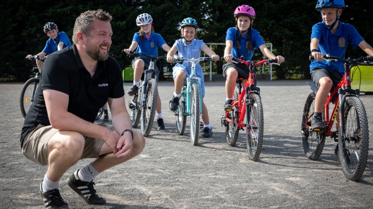 A male Walk Wheel Cycle Trust active travel officer crouches on the ground in school playground while a row of children on bikes wearing helmets line up beside him