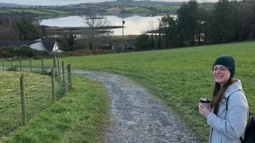 A woman wearing a hat and glasses turns to smile at the camera while walking along a country path with a lake in the background.