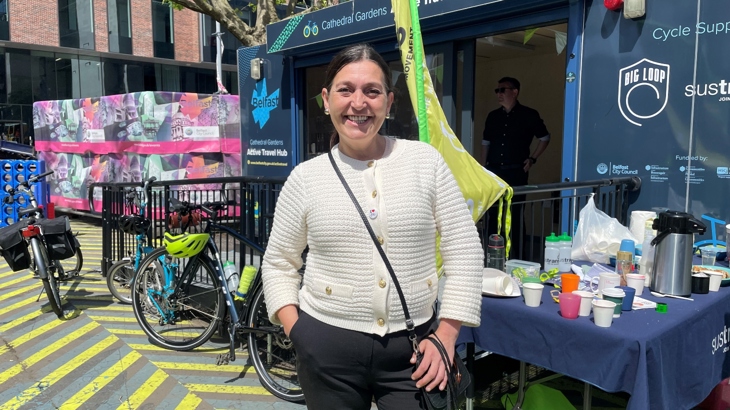 A woman with dark hair wearing an 'I heart cycling' badge on her cream cardigan smiles for the camera outside Cathedral Gardens hub.